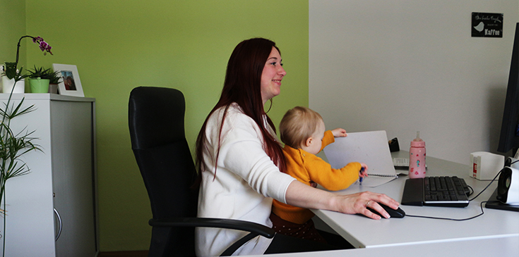 Eine Frau mit langen Haaren sitzt an ihrem Schreibtisch und arbeitet am PC. Auf ihrem Schoß sitzt ein Baby, das nach einem Block greift.