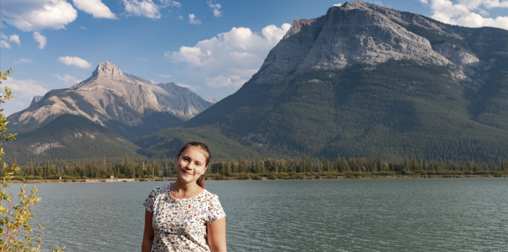 Eine junge Frau steht lächelnd vor einem See mit Bergen im Hintergrund.