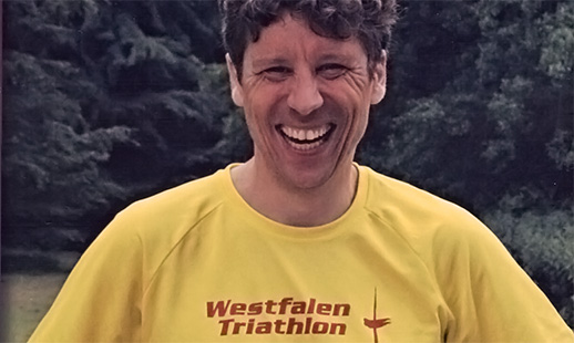 Ein Mann mit braunen Haaren, Martin Hörstemeier, steht lachend vor einem Wald. Er trägt ein gelbes T-Shirt mit der roten Aufschrift: „Westfalen Triathlon“