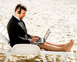 Ein Geschäftsmann sitzt mit Hemd, Jackett und Badehose im Strand und arbeitet.