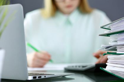 Eine Frau in hellgrüner Bluse sitzt an einem Schreibtisch mit aufgeklapptem Laptop, vor ihr liegen Aktenordner und ein Taschenrechner. Ihre Silhouette ist verschwommen und sie hält einen Stift in der Hand.