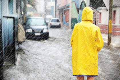 Person von hinten in gelbem Regenmantel steht in strömendem Regen auf einer überschwemmten Straße und schaut auf Häuser und Autos.