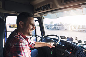 Ein Mann in kariertem Hemd sitzt am Lenkrad eines großen LKW. Er schaut aus der Frontscheibe heraus und lächelt.