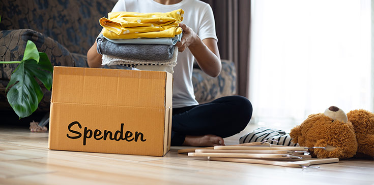 Eine Frau in T-Shirt und Jeans sitzt im Schneidersitz in ihrem Wohnzimmer und legt gefaltete Kleidung in eine Box mit der Aufschrift „Spenden“. Neben ihr liegen Kleiderbügel und ein Teddybär. Im Hintergrund sind eine Couch und eine Pflanze platziert.