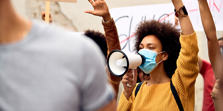 Eine Frau protestiert und ist umgeben von anderen Menschen, die mit ihr streiken. Sie trägt eine Maske, hat den einen Arm nach oben gestreckt und hält ein Megafon vor ihren Mund.