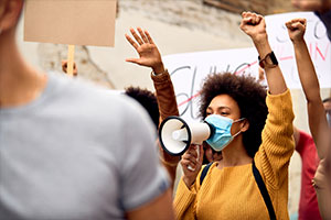 Eine Frau protestiert und ist umgeben von anderen Menschen, die mit ihr streiken. Sie trägt eine Maske, hat den einen Arm nach oben gestreckt und hält ein Megafon vor ihren Mund.