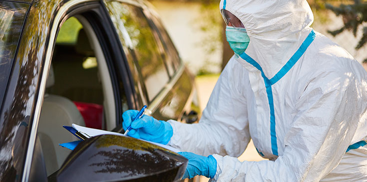 Eine Person in Schutzanzug steht am Autofenster und nimmt die persönlichen Daten auf.