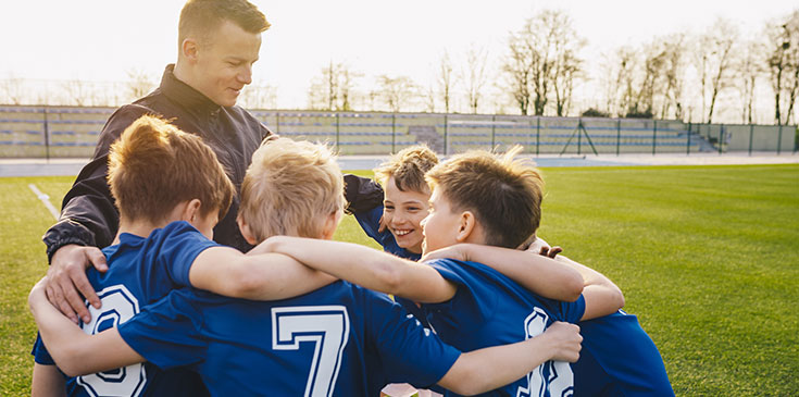 Ein Trainer und sein Team aus Jungs stehen im Kreis und haben die Arme über die Schultern der anderen gelegt. Alle lächeln.