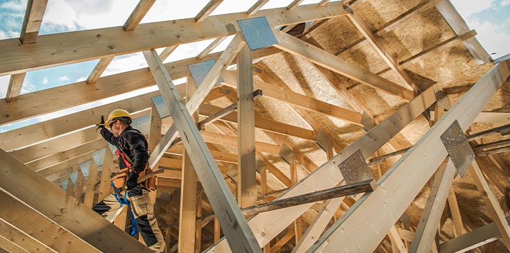 Ein Bauarbeiter mit Helm und Werkzeug steht auf einem Holzdach, das gerade ausgebaut wird. Durch das Dach hindurch sieht man den blauen Himmel