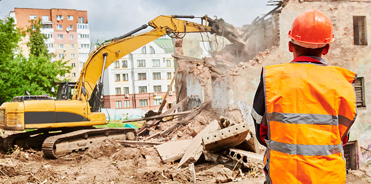 Ein Bauarbeiter steht mit dem Rücken zur Kamera und beobachtet, wie ein Bagger ein Haus abreißt.