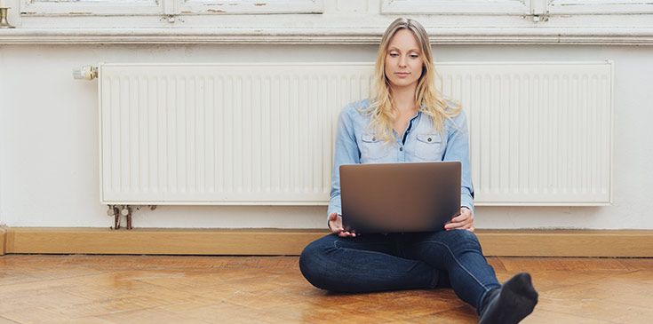 Eine junge Frau sitzt auf dem Boden vor der Heizung und schaut auf ihren Laptop.