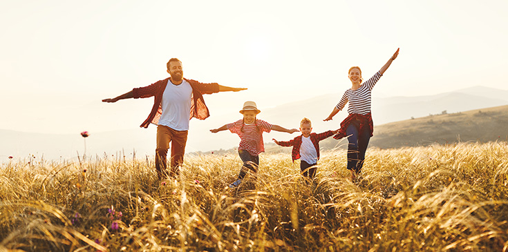 Eine Familie bestehend aus Mutter, Vater, Tochter und Sohn springt lachend über ein Feld während alle vier ihre Arme von ihrem Körper weg ausbreiten.