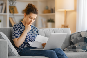 Eine junge Frau prüft ihren Steuerbescheid. Sie sitzt auf dem Sofa und hat ihren Laptop auf ihren Beinen.