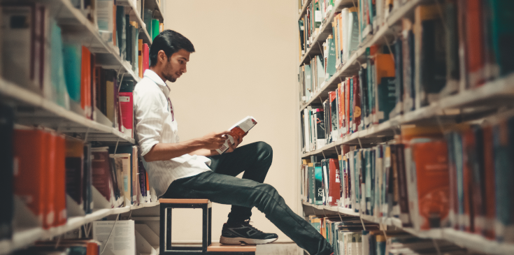 Ein junger Student ist in der Universitätsbibliothek und sitzt zwischen zwei großen Bücherreihen auf einem kleinen Tritt. Er trägt Jeans und Hemd und hält ein aufgeschlagenes Buch in der Hand, das er liest. Der Student ist von der Seite abgebildet, die Bücherreihen sind im Vordergrund verschwommen.