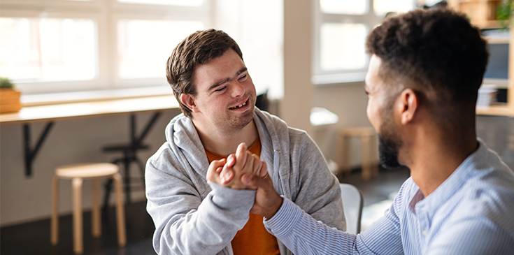 Ein junger Mann mit Down-Syndrom und ein anderer Mann sitzen sich lächelnd gegenüber in einem hellen Raum. Sie halten ihre Hände in einer freundschaftlichen Geste.