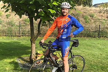 Ein Mann auf dem Fahrrad in Fahrrad-Dress und mit Helm lächelt in die Kamera. Im Hintergrund befindet sich ein Baum.