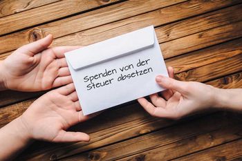 Rechts im Bild reicht eine Hand einen Briefumschlag mit der Aufschrift „Spenden von der Steuer absetzen“ an zwei Hände links im Bild weiter. Diese Hände sind offen gehalten, sodass der Umschlag leicht abgelegt werden kann.