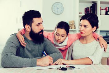 Ein Kind klammert sich zwischen seine Eltern, die sich anschauen. Der Vater hat einen Stift in der Hand, die Mutter zeigt mit dem Finger auf Papiere, die auf einem Tisch liegen.