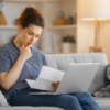 Eine junge Frau prüft ihren Steuerbescheid. Sie sitzt auf dem Sofa und hat ihren Laptop auf ihren Beinen.