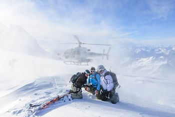 Drei Personen in Ski-Montur sitzen auf einem schneebedeckten Berg und schauen auf eine Karte. Im Intergrund sind weitere schneebedeckte Berge, blauer Himmel und ein Helikopter zu sehen, der Schnee aufwirbelt.