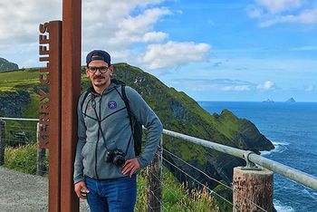 Ein Mann steht an einem Gerüst, man hat Blick auf Klippen und das Meer. Der Mann hat eine Mütze auf und sich eine Kamera umgehängt.