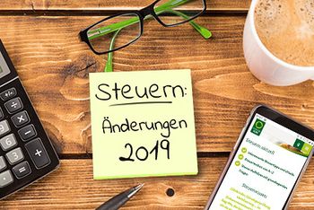 Ein gelber Notizzettel klebt auf einem Holztisch, darauf steht „Steuern: Änderungen 2019“. Um den Notizzettel herum liegen ein Taschenrechner, ein Kugelschreiber, eine Lesebrille und ein Smartphone mit der geöffneten Webseite des Steuerrings. Eine Tasse Milchkaffee rundet das Bild ab.