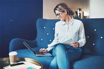 Eine Frau in Jeans und Bluse sitzt mit überschlagenen Beinen auf einem Sofa. Ihr Kopf ist nach rechts zu ihrem Laptop gedreht, den sie mit der rechten Hand bedient. Mit der anderen Hand hält sie einen Stapel Blätter auf ihrem Schoß. Vor ihr sind, auf einem kleinen Tisch, ein Coffee-to-go Becher sowie weitere Unterlagen platziert.