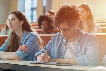 In einem Vorlesungsaal sitzen Studenten, dabei sind zwei deutlich zu erkennen. Eine Studentin schaut nach vorne, ein anderer Student schreibt etwas auf. Der Hintergrund ist verschwommen.