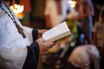 Ein Priester im hellen Gewand ist von der Seite zu sehen. Er hält die Bibel aufgeschlagen in seiner Hand. Im Hintergrund findet ein Gottesdienst statt.