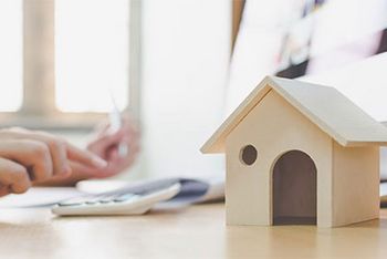 Im Vordergrund des Bildes steht ein kleines Modell-Holzhaus. Der Hintergrund ist verschwommen, mit der rechten Hand tippt jemand auf einem Taschenrechner und in der linken Hand hält die Person einen Stift.