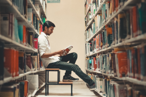 Ein junger Student ist in der Universitätsbibliothek und sitzt zwischen zwei großen Bücherreihen auf einem kleinen Tritt. Er trägt Jeans und Hemd und hält ein aufgeschlagenes Buch in der Hand, das er liest. Der Student ist von der Seite abgebildet, die Bücherreihen sind im Vordergrund verschwommen.