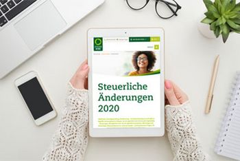 Zwei Frauenhände halten ein Tablet. Auf dessen Bildschirm ist die Steuerring-Website und die Aufschrift „Steuerliche Änderungen 2020“ zu erkennen. Um das Tablet herum liegen auf einem Schreibtisch eine Brille, ein Handy, ein Block mit Stift, eine Pflanze und ein Laptop.