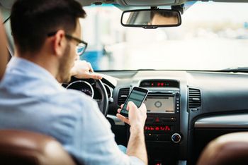 Ein Mann sitzt im Auto. Mit seiner linken Hand hält er das Steuer, in der rechten Hand hat er sein Handy. Man sieht den Rücken des Mannes und sein Profil.